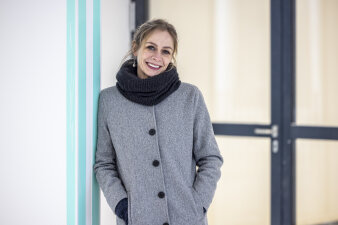 eine Frau mit hellgrauem Mantel und dunkelgrauem Schal lehnt an einer Wand
