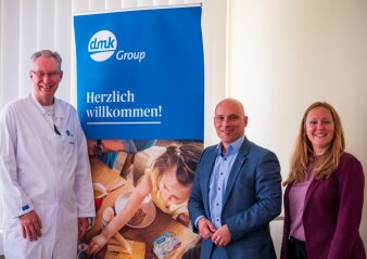 drei Personen, davon eine in weißem Kittel, stehen neben einem Roll-up mit Unternehmenswerbung für die dmk Group
