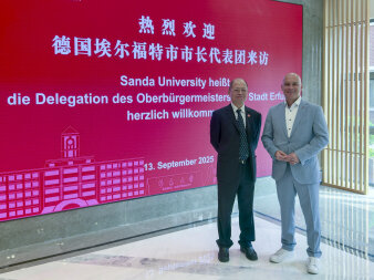 zwei Männer stehen vor einer Wandprojektion, in der auf rotem Hintergrund chinesisches und deutsche Schrift ist