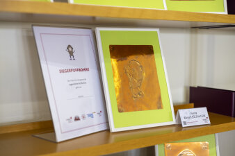 Auf einem Regal steht eine auf Goldpapier gekratzte Puffbohne. Dabei steht eine Urkunde.