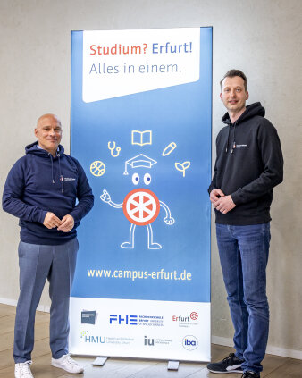 Zwei Männer stehen vor einem Plakat, das fürs Studium wirbt.