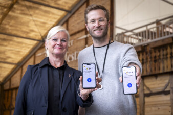 zwei Personen halten ein Handy in die Kamera, auf dem Display ist der Schriftzug "Drücken um Helfer alarmieren"