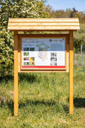 Eine Informationstafel auf einer Wiese, im Hintergrund Bäume