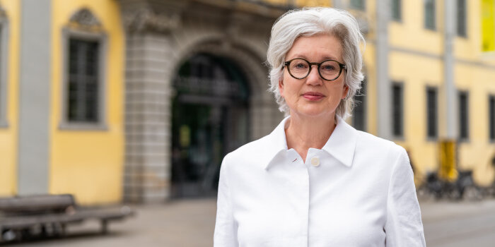 Eine Frau mit weißgrauen kurzen Haaren, einer runden dunklen Brille und weißer Bluse vor einem Gebäude