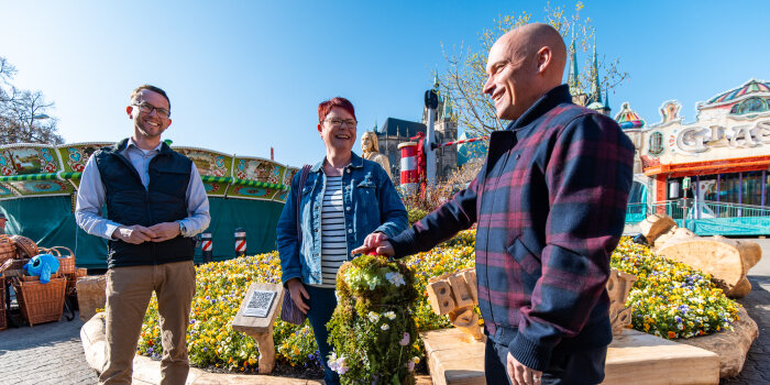 Drei Personen stehen vor einem Blumenbeet und drücken einen Startknopf