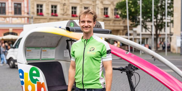 Ein Mann steht vor einem überdachtem Lastenrad auf einem Platz umgeben von historischen Gebäuden