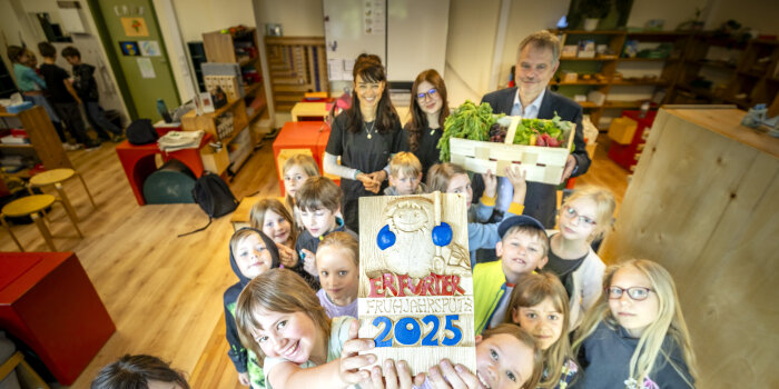 Eine Kindergruppe steht in einem Klassenzimmer und hält einen Holzpokal in die Luft