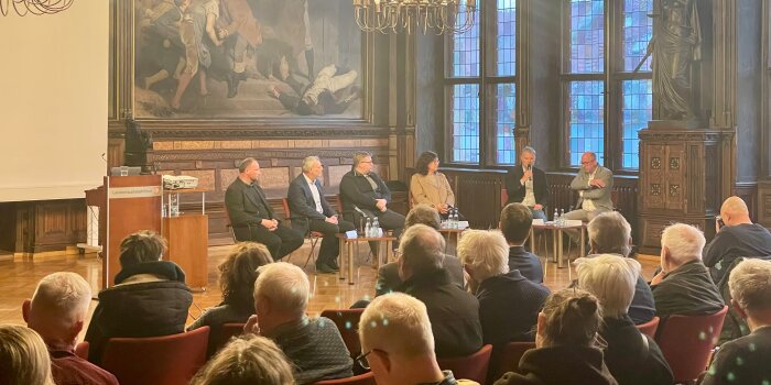 Menschen sitzen in einem historischen Saal mit großen Fenstern, im Podium sitzen Personen in einem Halbkreis