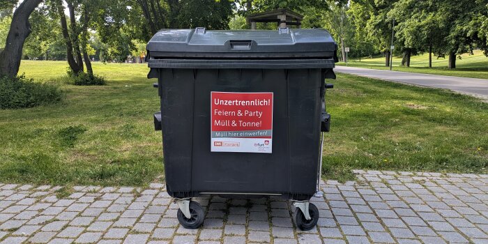 Ein großer schwarzer Müllcontainer steht an einem Parkweg umgeben von grünen Bäumen