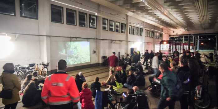 Eine Gruppe von Personen steht mit Fahrrädern in einer Halle und blickt auf eine Leinwand