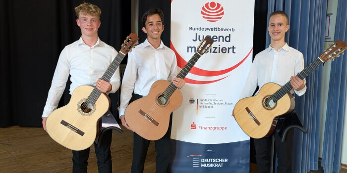 Drei junge Männer mit Gitarre stehen beisammen vor einem Plakat von „Jugend musiziert“.