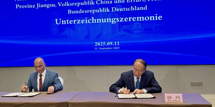 zwei Männer sitzen an einem Tisch unter unterschreiben ein Schriftstück, im Hintergrund eine Projektion mit Schrift