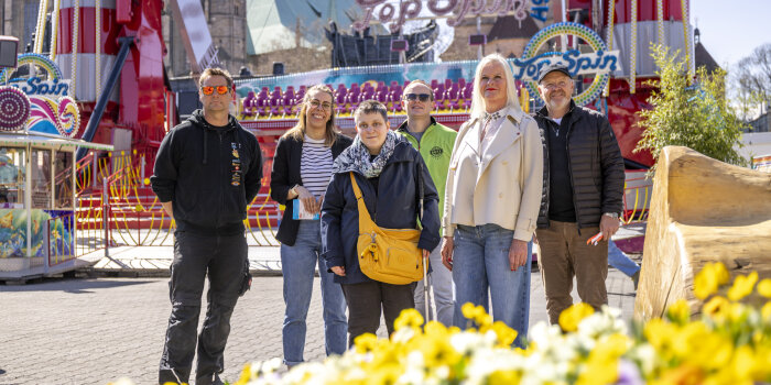Eine Gruppe von Personen steht auf einem Volksfest vor einem Fahrgeschäft, im Vordergrund Frühlingsblumen