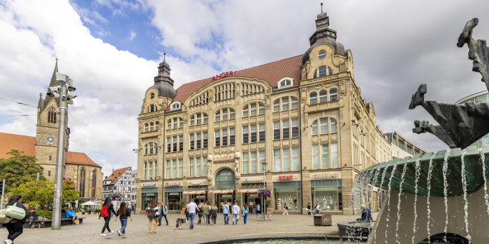 ein fünfstöckiges Gebäude mit Schaufenstern im Erdgeschoss, im Vordergrund ein Springbrunnen