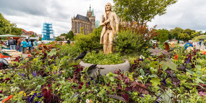 Ein bunt bepflanztes Blumenbeet, in der Mitte eine Holzskulptur und im Hintergrund der Dom