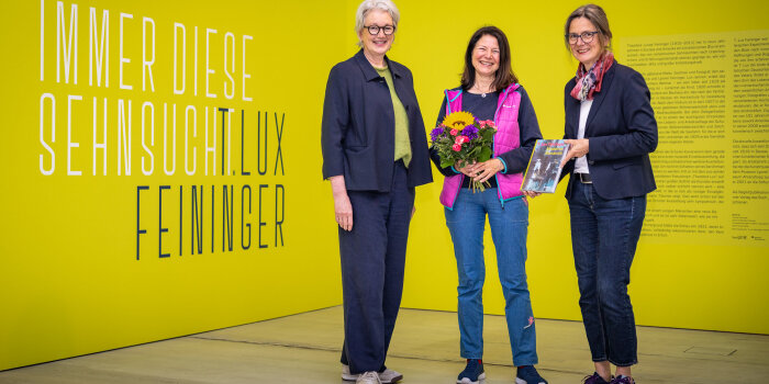 Drei Frauen stehen beisammen, die mittlere hält einen Blumenstrauß. Sie stehen vor einer gelben Wand mit Aufschrift