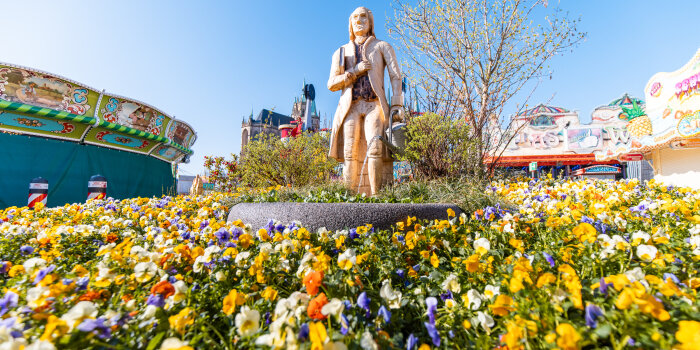 Ein Blumenbeet bepflanzt mit Frühjahrsblühern, in der Mitte steht eine hölzerne Statue