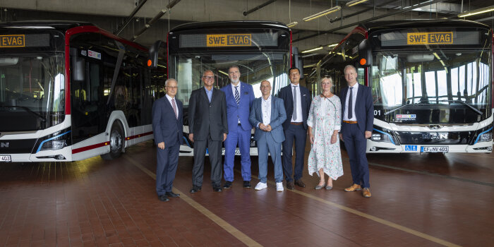sieben Personen stehen vor drei Bussen in einer Halle
