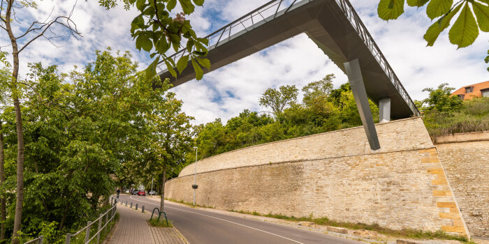 Eine Festungsmauer an einer Straße. Darüber hinweg verläuft eine eiserne Brücke. 