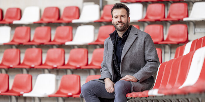 ein Mann im grauen Mantel sitzt auf einer Stadiontribüne mit roten und weißen Schalensitzen