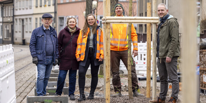 mehrere Personen stehen an einem frisch gepflanzten Baum in urbaner Umgebung