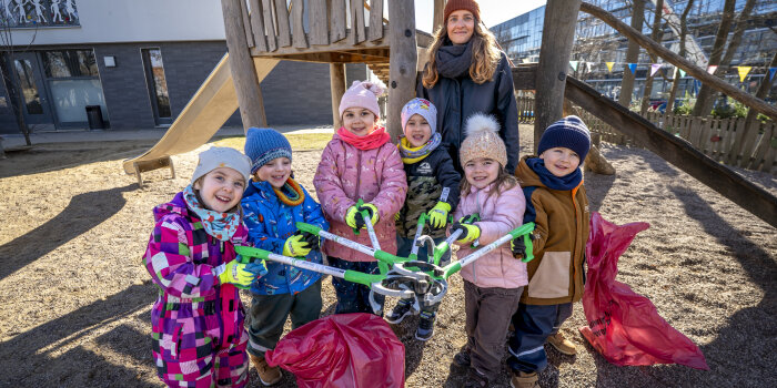 Kinder stehen mit einer erwachsenen Person auf einem Spielplatz und halten Müllgreifer und -säcke in der Hand
