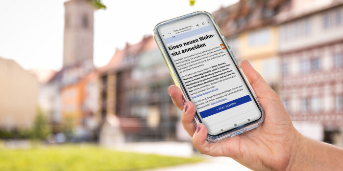 Ein Handy zeigt die Website der elektronischen Wohnsitzanmeldung, dahinter die Krämerbrücke