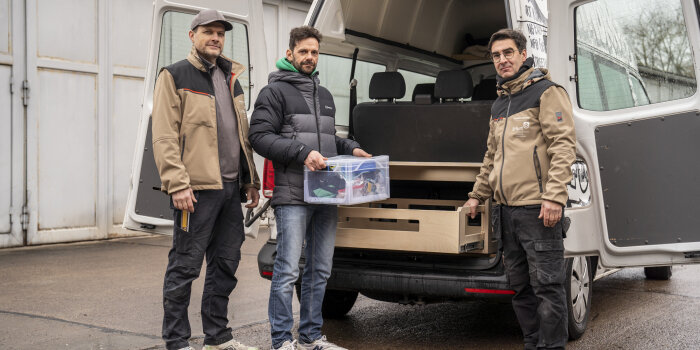 Drei Männer stehen vor einem Bus mit offenen Hecktüren. Der Mittlere hält eine Kiste in der Hand. 