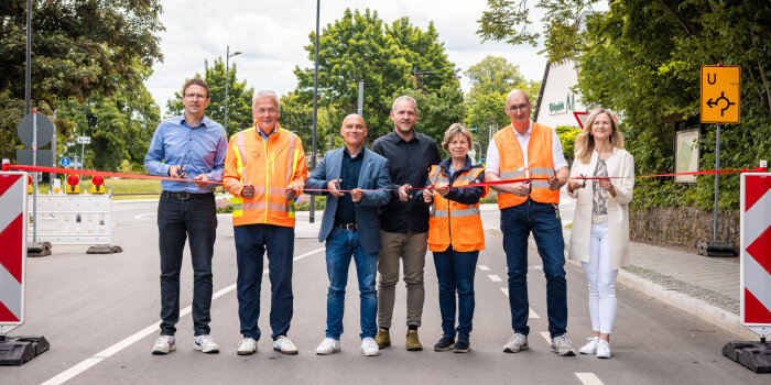 Fünf Männer und zwei Frauen stehen auf einer Straße und schneiden ein rotes Band durch. 