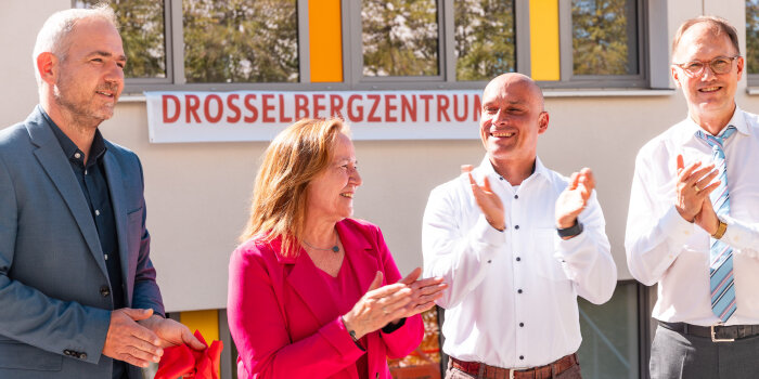 Ein saniertes Haus. Im Vordergrund drei Männer und eine Frau, die ein Band zerschneiden und klatschen.  