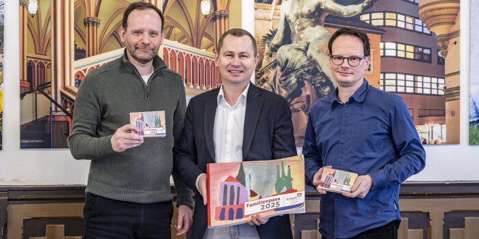 Drei Männer stehen. Der linke und rechte halten je ein kleines Heft in der Hand, der Mittlere ein großes Exemplar. 