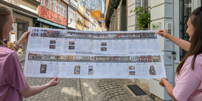 Zwei Frauen stehen zwischen historischen Gebäuden und halten eine Zeitung in der Hand
