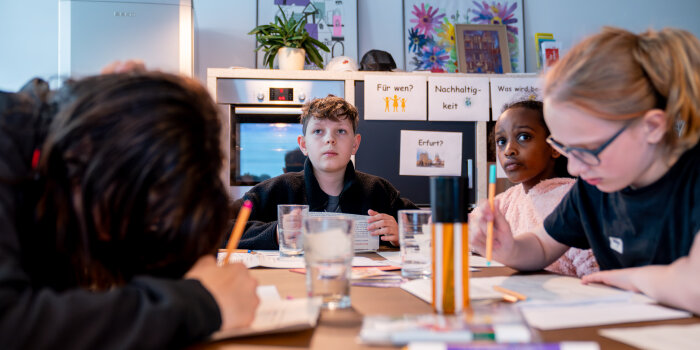 Vier Kinder sitzen an einem Tisch. Drei blicken auf und eines hat den Kopf gesenkt. 