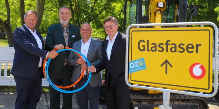 vier Männer halten gemeinsam zwei in Rollen gelegt Kabel, im Vordergrund ein Ortsschild mit Schriftzug "Glasfaser"