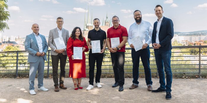 sieben Personen stehen in einer Reihe, fünf von ihnen halten eine Urkunde in der Hand, Erfurter Dom im Hintergrund