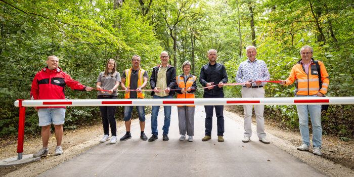 Mehrere Personen stehen auf einer Laufbahn im Wald hinter einer Schranke und schneiden ein Band durch.