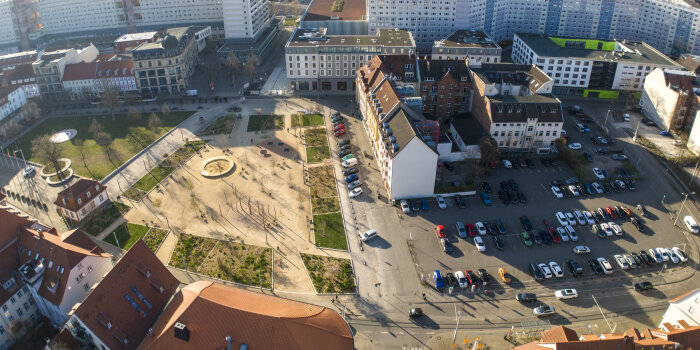 Luftaufnahme von einer Grünfläche mit Spielplatz und Parkplatz, dazwischen eine Straße mit parkenden Fahrzeugen