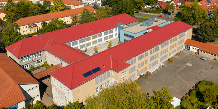 Luftbild einer Schule mit rotem Satteldach und zwei Solarmodulen.