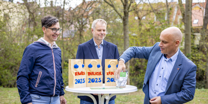 Drei Männer an einem weißen Stehtsich, darauf Holzpokale. Ein Mann greift in eine Glasschale und zieh ein Los 