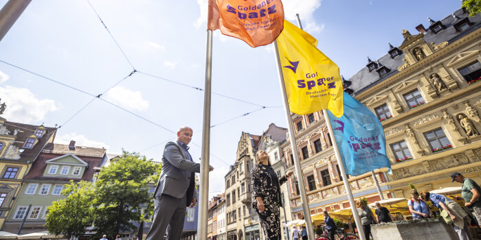 Foto: Flagge für den Goldenen Spatz wird gehisst © Stadtverwaltung Erfurt Zwei Personen hissen drei Flaggen vor dem Erfurter Rathaus