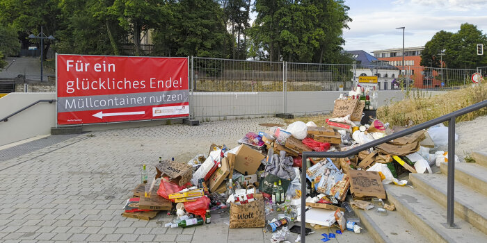 ein Haufen Müll liegt am Fuße einer Treppe, im Hintergrund eine Bauzaunplane mit Aufschrift "Müllcontainer nutzen"