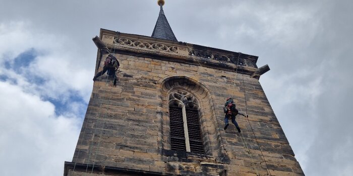 Zwei Männer mit Helm und Kletterausrüstung hängen an einem Turm.