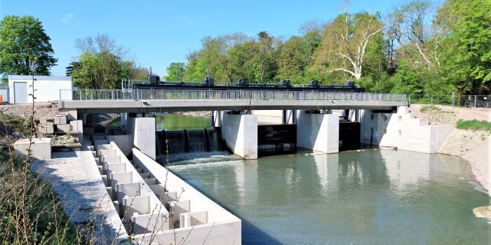 Eine Brücke und Wehr über einem Flusslaif umgeben von grünen Bäumen und blauem Himmel