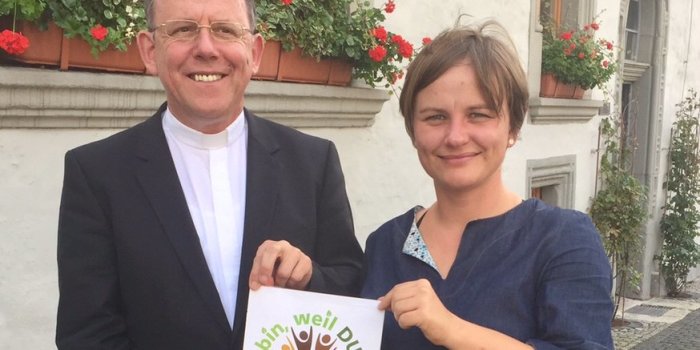 Frau und Mann mit einem Blatt in den Händen mit Text: "Ich bin, weil du bist"