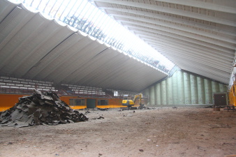 Ein Kleinbagger in der 60 Meter langen Sporthalle arbeitet im Tageslichlicht, das durch das Glasdach in die Halle fällt.