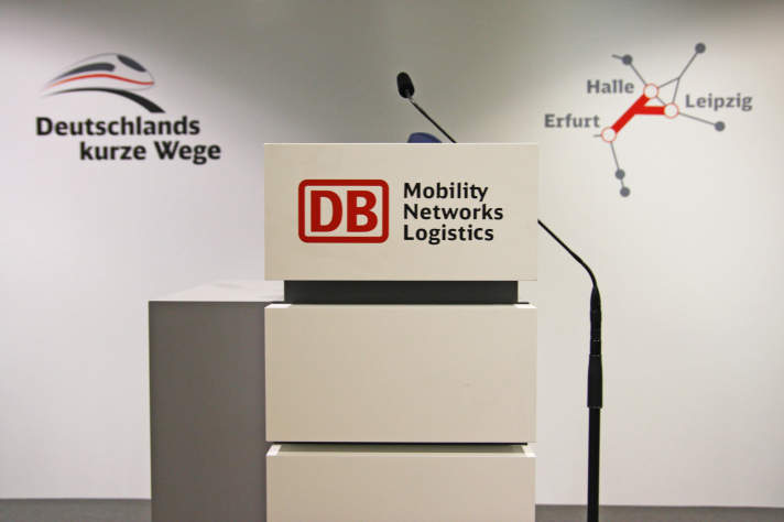 Rednerpult mit dem Deutschen Bahn Logo und Mikrofon vor einer weißen Wand, auf dessen weitere Logos bezüglich des Projektes zu sehen sind.