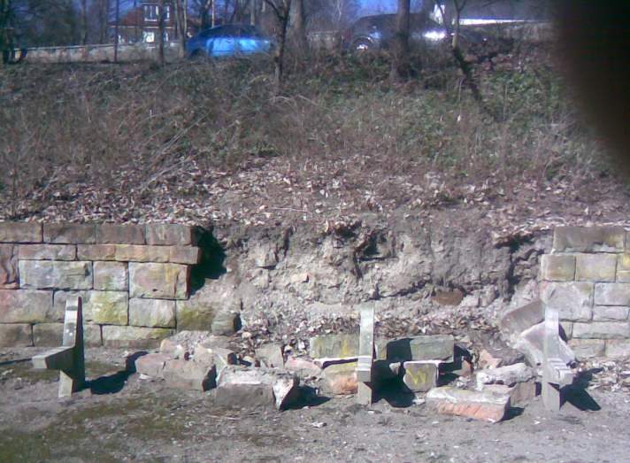 Eine Natursteinmauer ist zum Teil eingerissen, zum Teil steht sie noch. Darüber ein kleiner Hand mit Gras und Getrüpp.