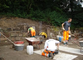 Drei Fachkräfte in Arbeitskleidung bauen eine Natursteinmauer. Um sie herum stehen Arbeitsmittel wie Schubkarre oder Wasserkanister.