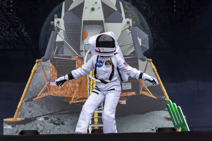 Ein als Astronaut verkleideter Darsteller steht vor einer Attrappe eines Raumschiffs auf dem Mond.