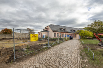 Weg mit Kopfsteinpflaster und Bauzaun führt zu einem länglichen historischen Gebäude auf Baustelle